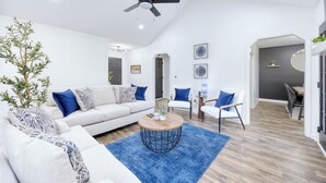 Una sala moderna con un sofá blanco, cojines estampados en azul y blanco, una mesa de centro redonda y un ventilador de techo.