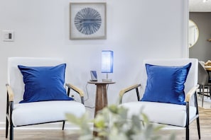 Una habitación moderna con dos sillones blancos, cada uno con un cojín azul, y una pequeña mesa de madera con una lámpara azul en el centro.