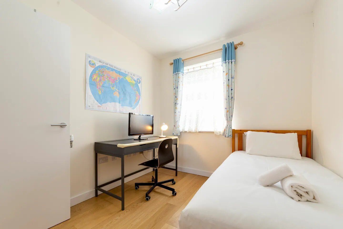 Ein Schlafzimmer mit einem Bett, einem Schreibtisch mit Computer, einem Stuhl, einer an der Wand hängenden Karte und einem Fenster mit Vorhängen.