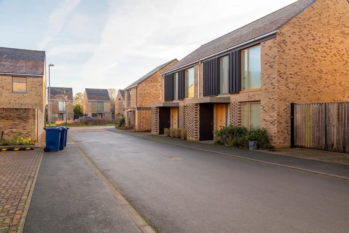 A residential street with modern brick houses, a paved road, and a blue trash bin.