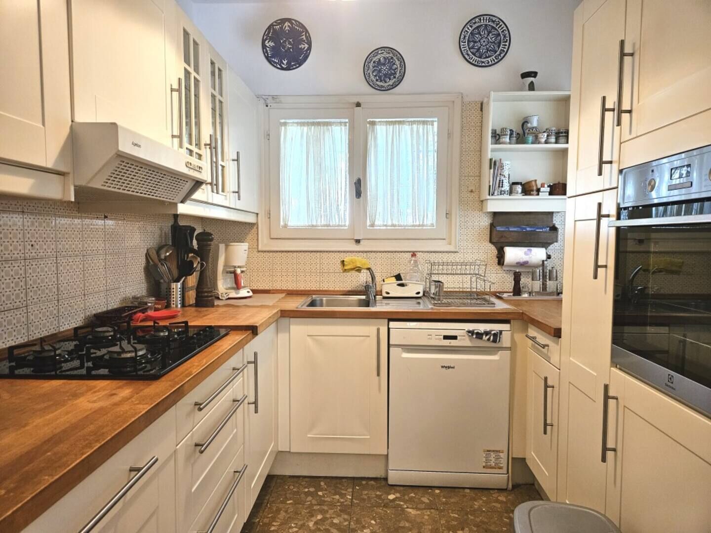Een compacte keuken met witte kastjes, een ingebouwde oven, een vaatwasser en een raam met gordijnen.