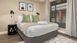 Un dormitorio ordenado con cama, almohadas, una mesita de noche y una ventana con persianas.