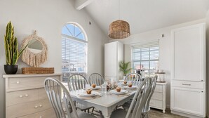A dining area with a white table set for four, surrounded by grey chairs. There is a wicker basket on the table, a potted plant, and a macrame wall hanging. - Walk to Beach, BBQ Patio Area, Community Pool, Games: Beach Soulcation (Galveston)