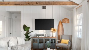 A living room with a large flat-screen TV mounted on the wall, a wooden ceiling beam, a striped armchair, a white sofa, and a potted plant. - Walk to Beach, BBQ Patio Area, Community Pool, Games: Beach Soulcation (Galveston)