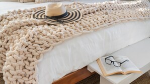 A bed with a chunky knit blanket, a straw hat, and an open book with glasses on a bedside table. - Walk to Beach, BBQ Patio Area, Community Pool, Games: Beach Soulcation (Galveston)