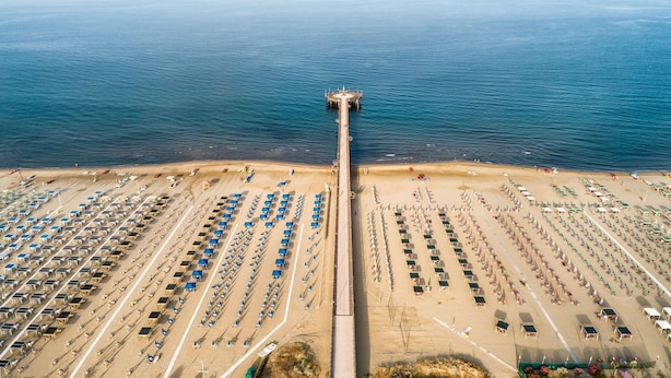 An aerial view of a beach with rows of sun loungers and umbrellas, a pier extending into the sea, and a clear sky.