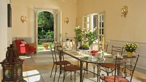 A dining area with a glass table, metal chairs, and a view of a garden through an open doorway.