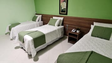A hotel room with three beds, each with white bedding and green accents. There is a small bedside table with a phone and a plant. The walls are painted green, and there is a framed picture on the wall.