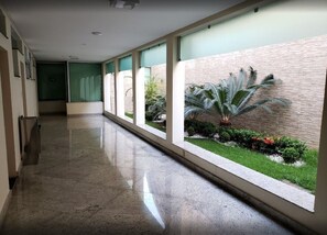 A long, narrow corridor with a polished floor, large windows, and a view of a landscaped garden with a palm tree. - Domus Cidade Nobre (Ipatinga)