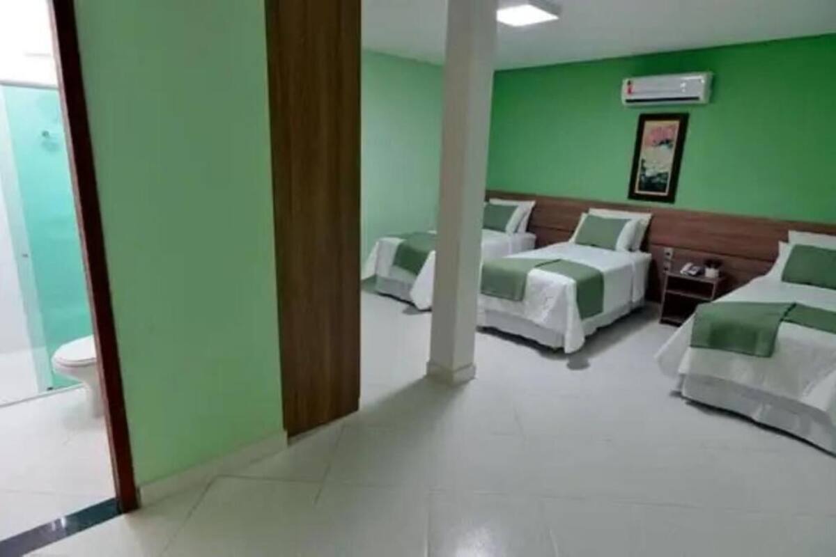 Quarto de hotel com três camas, parede verde, chão branco e banheiro à vista.