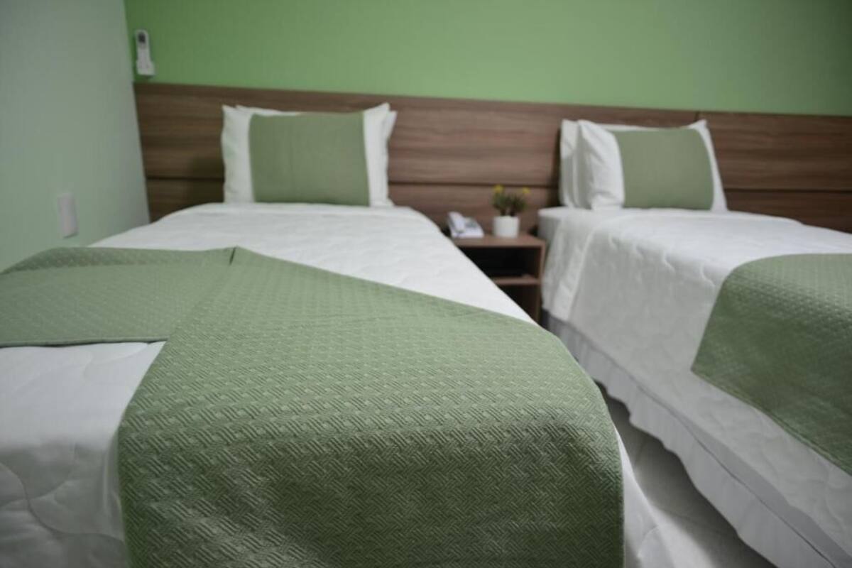 Quarto de hotel com duas camas, cada uma com um cobertor verde e roupa de cama branca. Há uma cabeceira de madeira, uma mesa de cabeceira com um abajur e uma pequena mesa com uma planta.