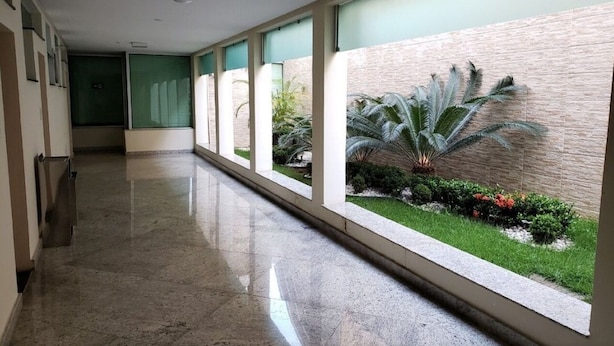 A long, narrow corridor with a polished floor, large windows, and a view of a landscaped garden with a palm tree. - Domus Cidade Nobre (Ipatinga)