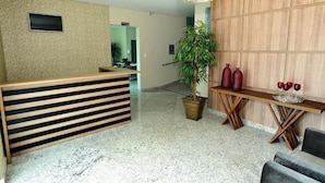 A reception area with a wooden reception desk, a potted plant, a small table with red vases, and a staircase in the background. - Domus Cidade Nobre (Ipatinga)