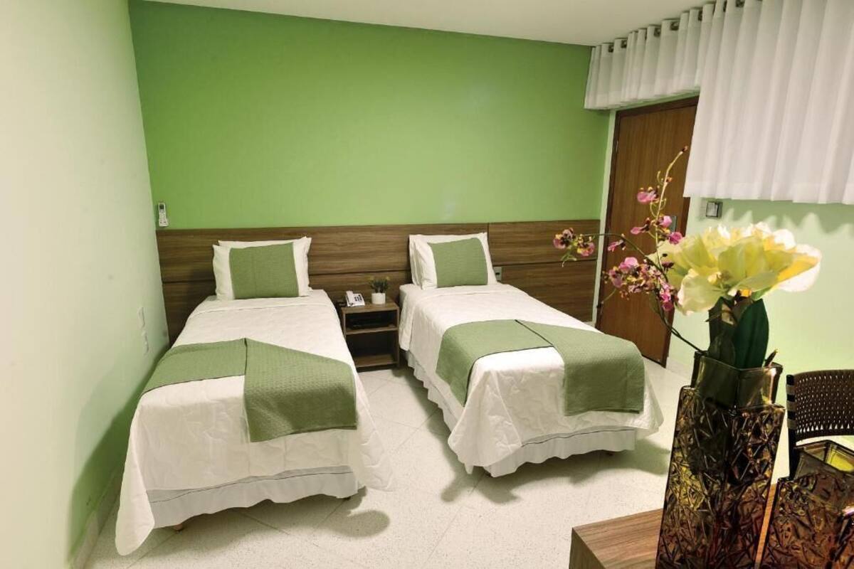 Quarto de hotel com duas camas de solteiro, uma mesinha de cabeceira, um vaso com flores e uma porta de madeira.