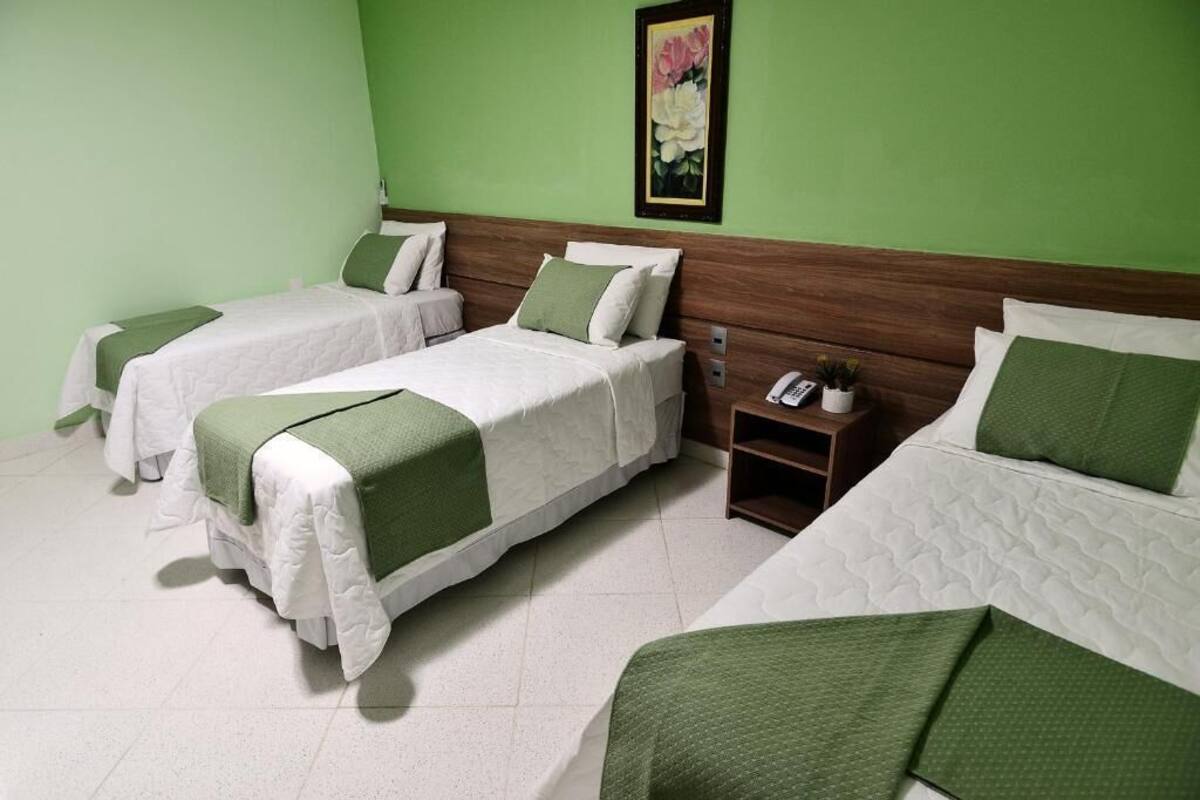 Quarto de hotel com três camas, cada uma com roupa de cama branca e detalhes verdes. Há uma pequena mesa de cabeceira com um telefone e uma planta. As paredes são pintadas de verde, e há um quadro pendurado na parede.
