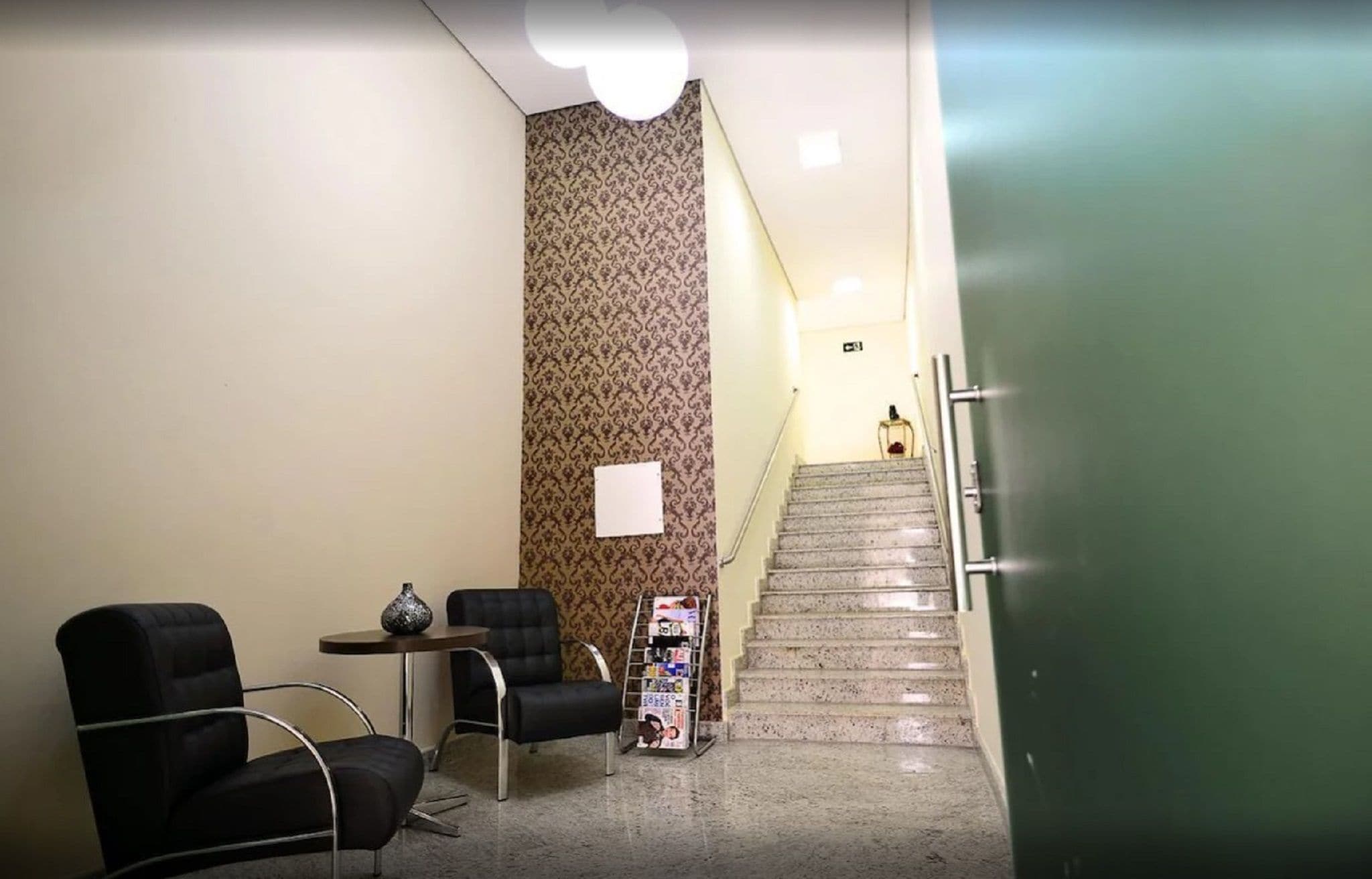A reception area with a staircase leading to an upper floor, a reception desk, two chairs, a small table with a vase, and a magazine rack.
