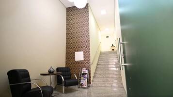 A reception area with a staircase leading to an upper floor, a reception desk, two chairs, a small table with a vase, and a magazine rack.