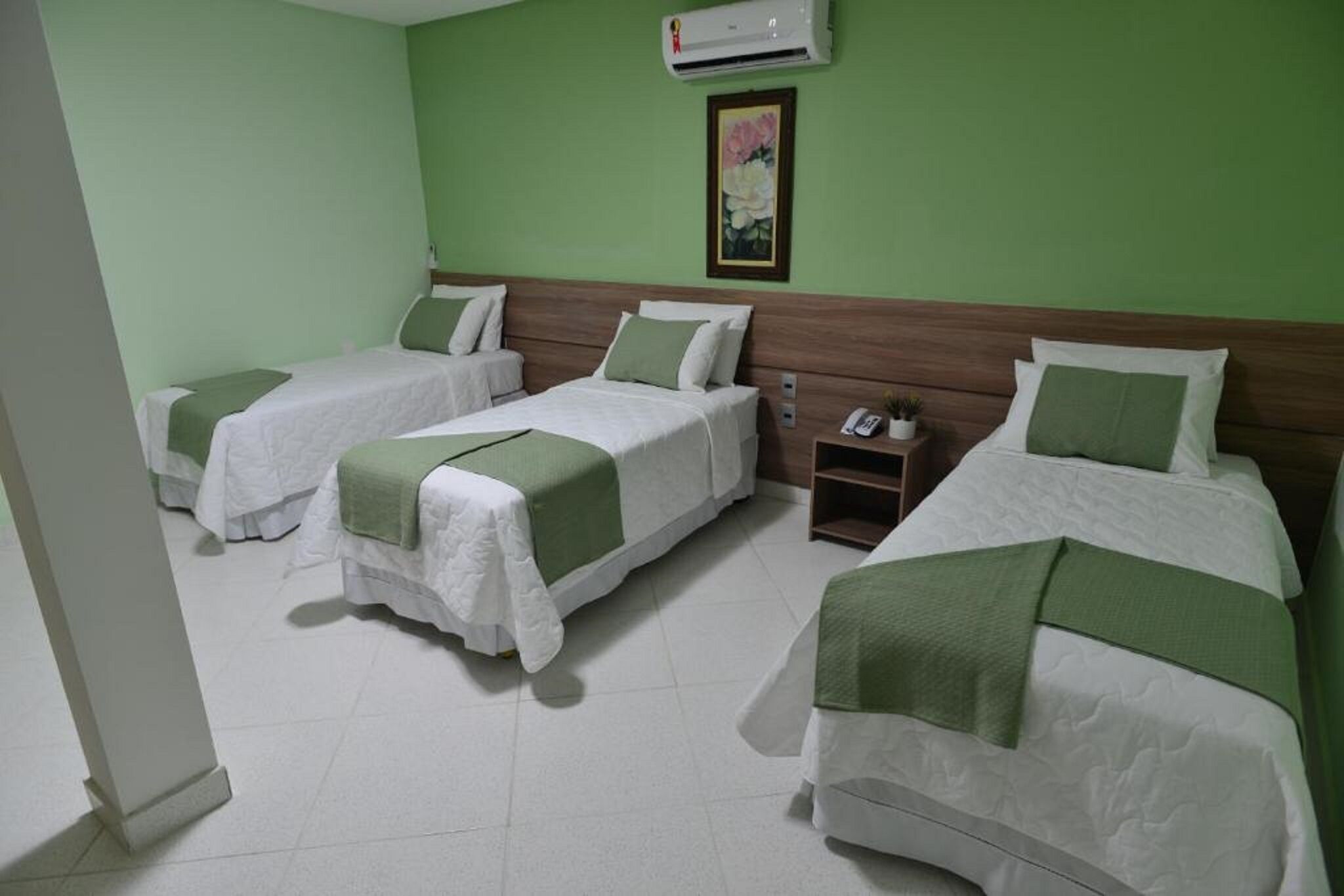 Quarto de hotel com três camas de solteiro, cada uma com colcha verde e almofadas brancas. Possui cabeceira de madeira, uma mesinha de cabeceira com abajur e ar-condicionado fixado na parede.