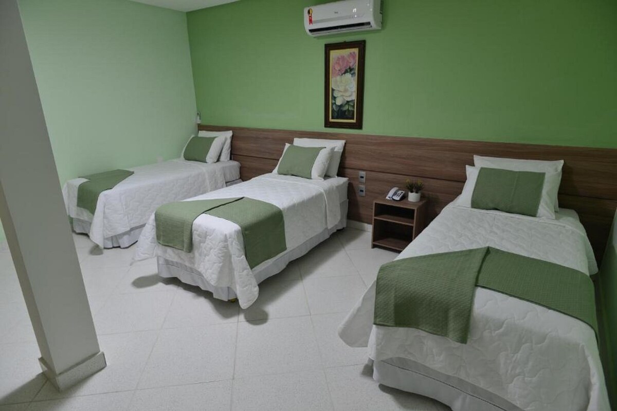 A hotel room with three single beds, each with a green bedspread and white pillows. There is a wooden headboard, a small bedside table with a lamp, and a wall-mounted air conditioner.