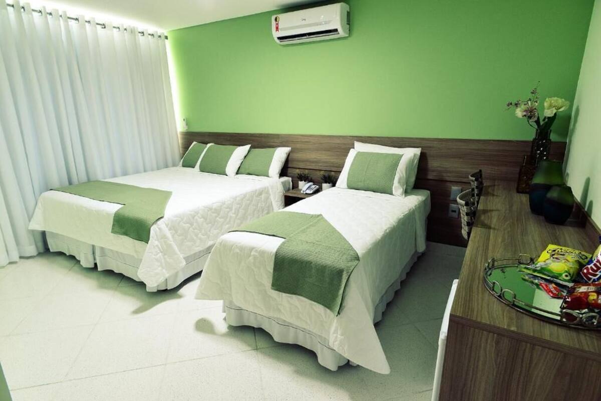 Quarto de hotel com duas camas, uma mesa com lanches, um vaso com flores e um ar condicionado na parede.
