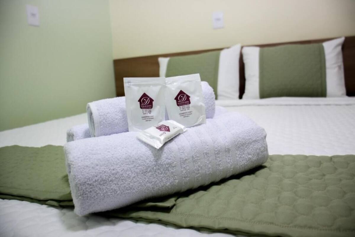 Cama bem arrumada com roupa de cama branca e verde, duas toalhas e dois artigos de higiene pessoal num quarto de hotel.