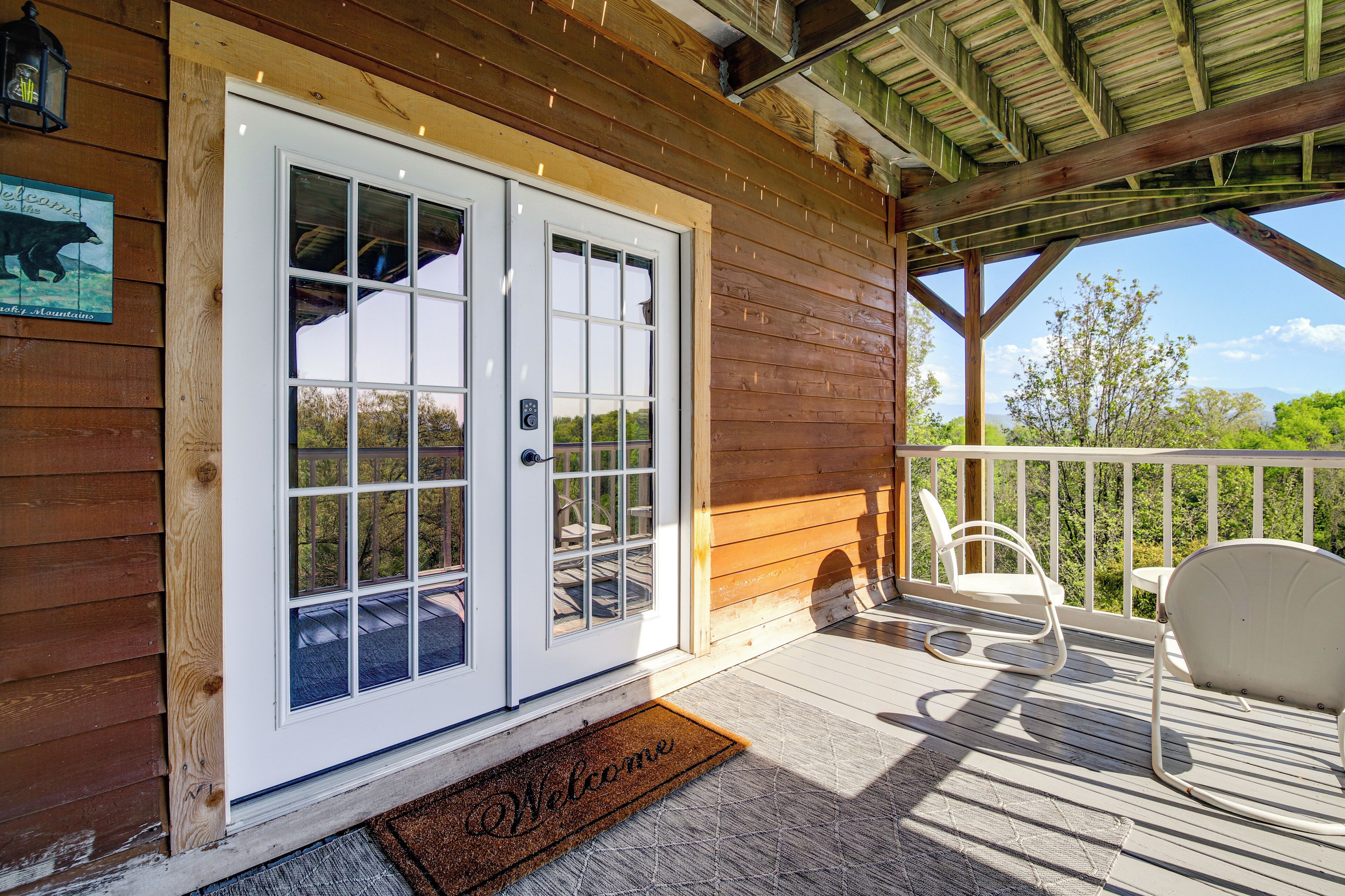 A wooden cabin with a white door, a porch with chairs, and a welcome mat.