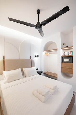 A modern hotel room with a large bed, a ceiling fan, a bedside table, and a built-in shelf with books and a safe. - Vaya Suites (Malevizi)