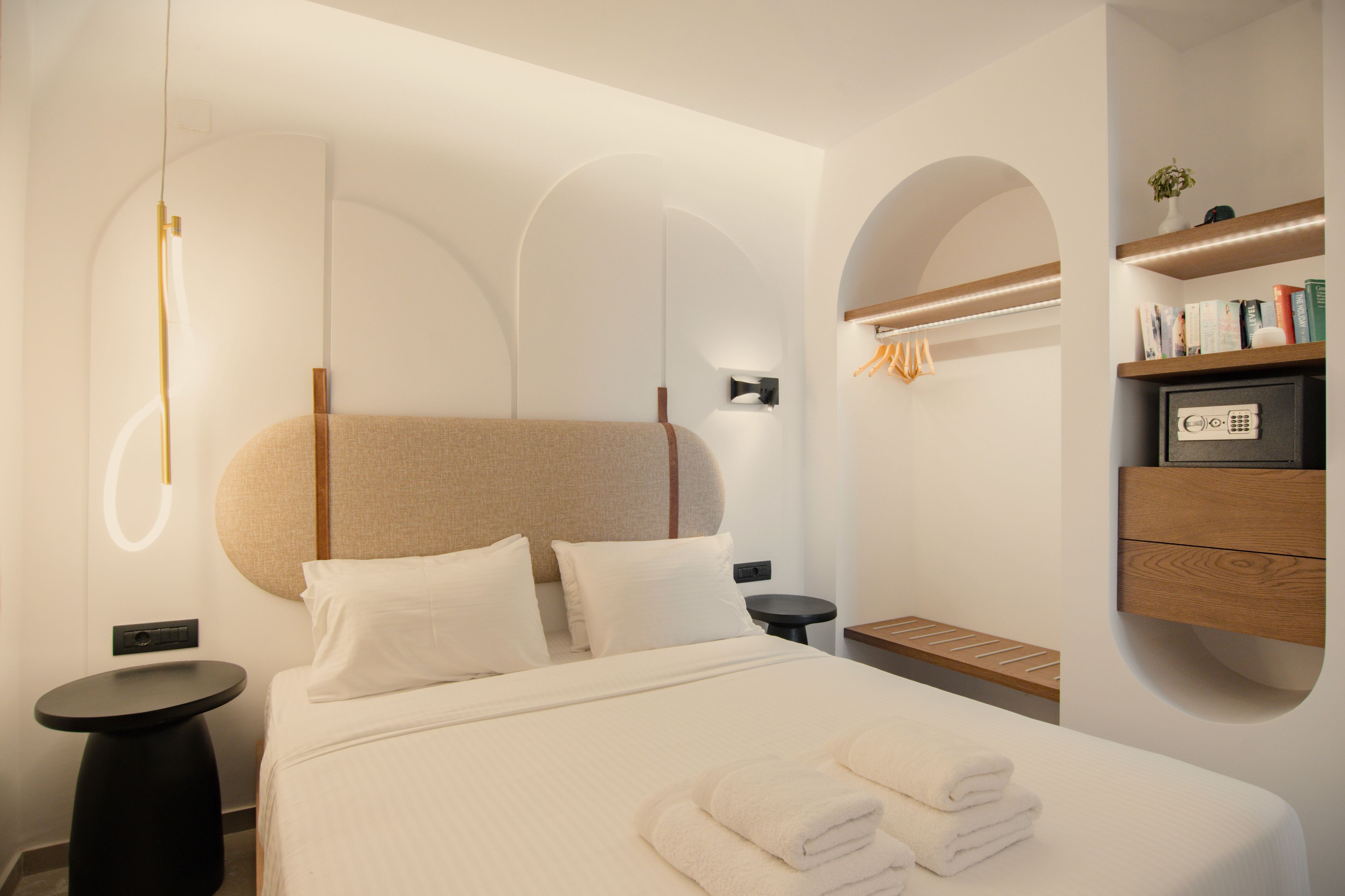 A modern hotel room with a large bed, a round headboard, a bedside table, a small round table, and a built-in shelf with books and a vase.