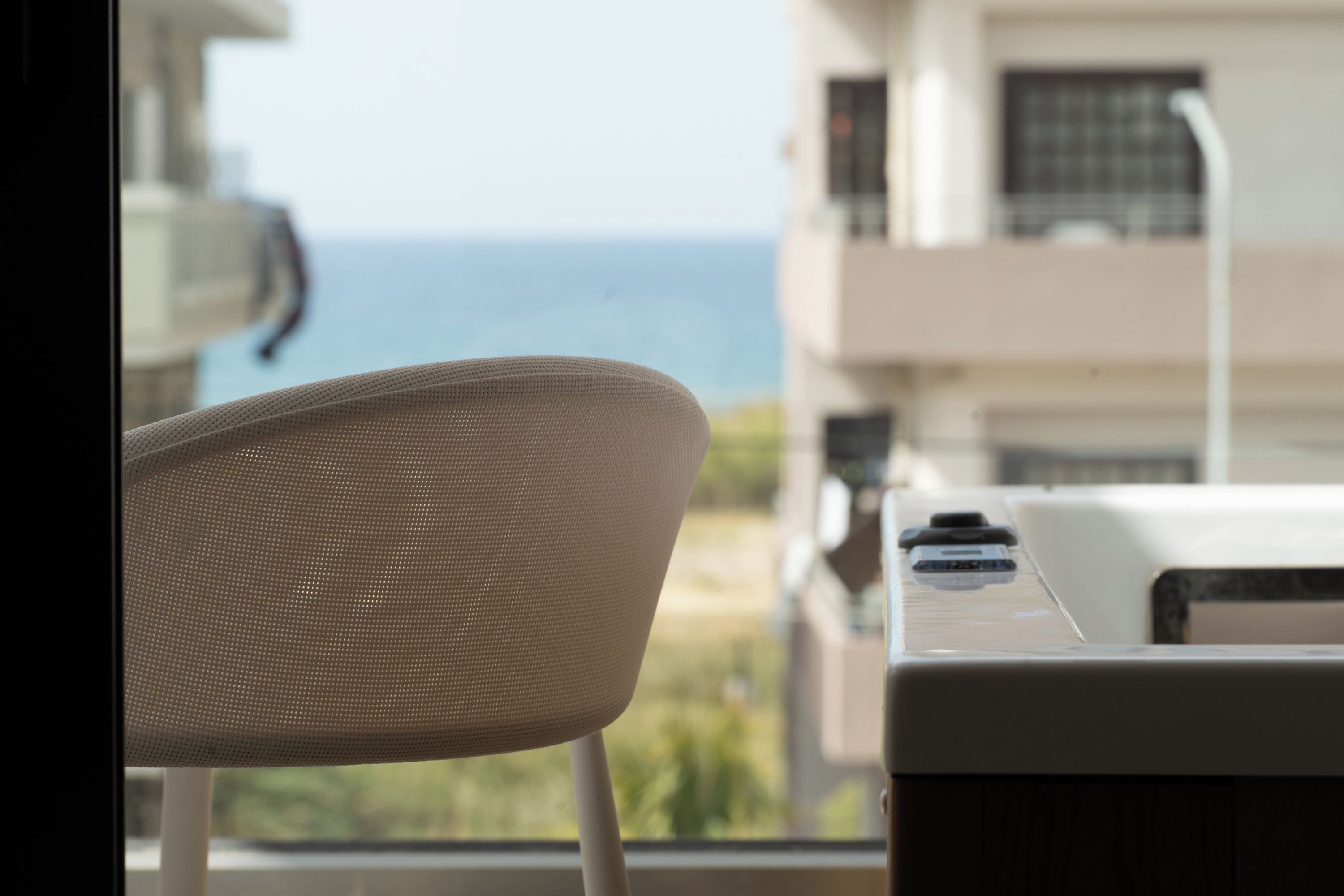 Una silla con respaldo de malla, una ventana con vista al mar y un edificio con balcones.