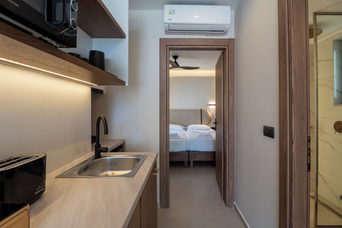 A compact kitchen with a toaster, sink, and microwave, leading to a bedroom with a bed and ceiling fan.