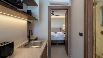 Cocina compacta con tostadora, lavamanos y microondas, que da paso a un dormitorio con cama y ventilador de techo.