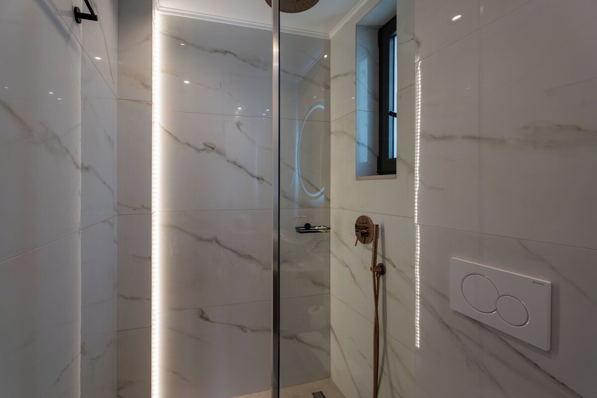 A modern bathroom with a glass shower enclosure, a handheld showerhead, and a wall-mounted control panel.