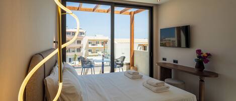 A modern hotel room with a large bed, a desk, a TV, and a balcony with outdoor seating.