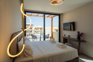 A modern hotel room with a large bed, a desk, a TV, and a balcony with outdoor seating. - Vaya Suites (Malevizi)