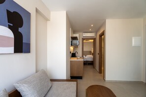 Een moderne hotelkamer met een bank, een klein houten tafeltje, een kitchenette en een gang naar een slaapkamer.