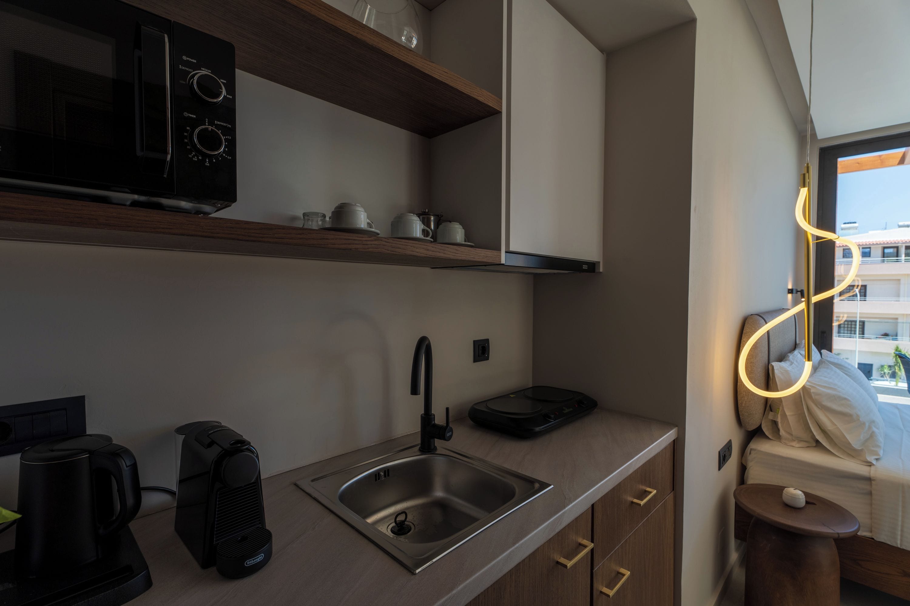 Una kitchenette moderna equipada con microondas, cafetera y hervidor, un lavamanos de acero inoxidable y una pequeña mesa con lámpara.