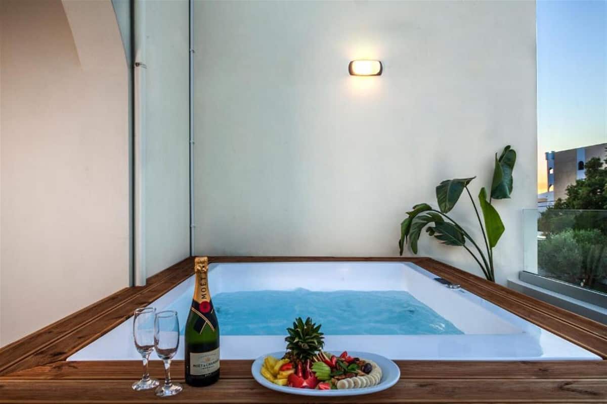 A hot tub with a wooden deck, a bottle of champagne, two glasses, and a plate of fruit.