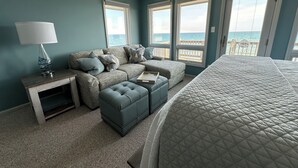 Un dormitorio con una cama grande, un sofá, un reposapiés y una mesita de noche. Hay ventanas amplias que dan a vistas del océano.
