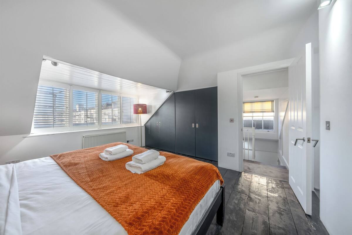 A modern bedroom with a large bed, a window with blinds, a dark wardrobe, and a door leading to another room.
