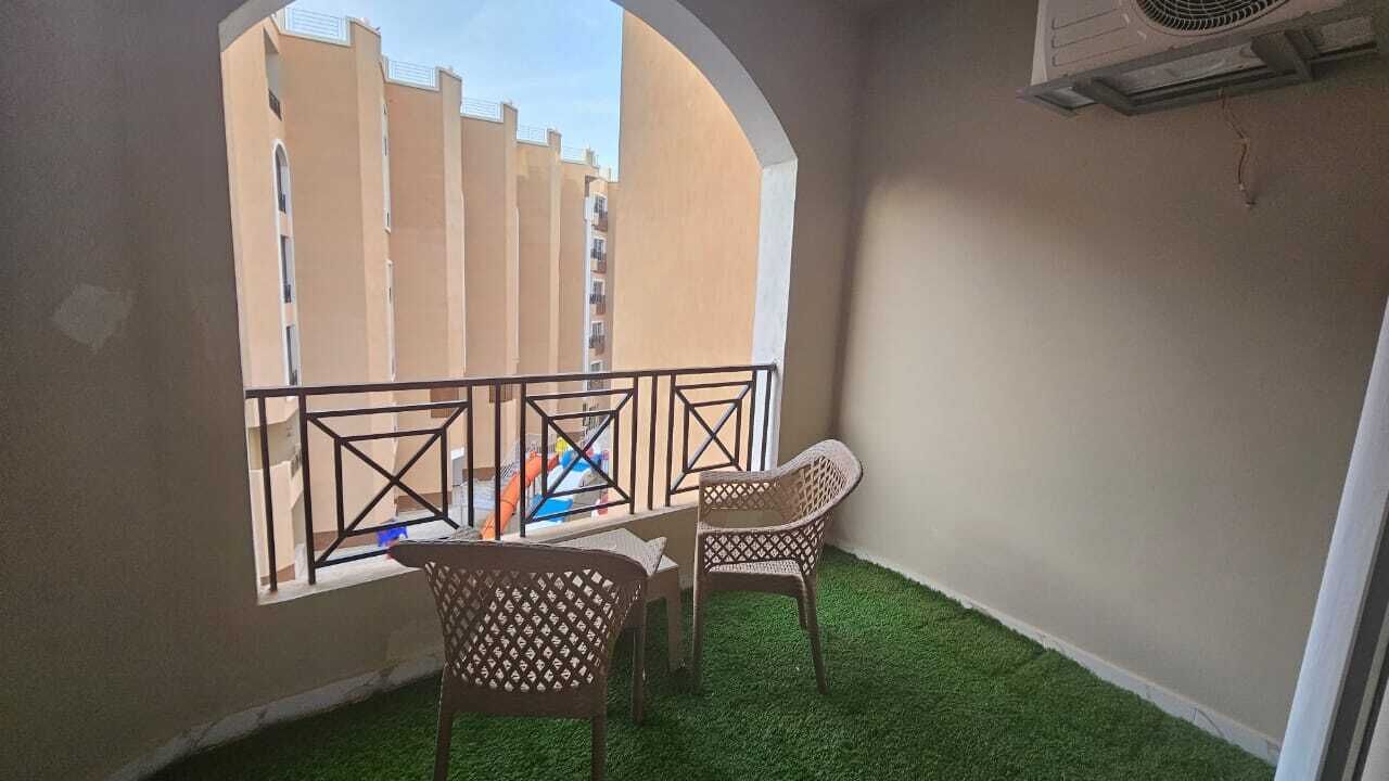 Un balcon donnant sur des immeubles, deux fauteuils en osier et un appareil de climatisation.