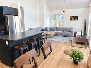 A modern kitchen with a central island, dining area with wooden chairs, a grey sofa, and a small table with a plant. - Modern Spa Chalet near Lac des Îles · Ski Montcalm 20 min · Pets OK (Entrelacs)