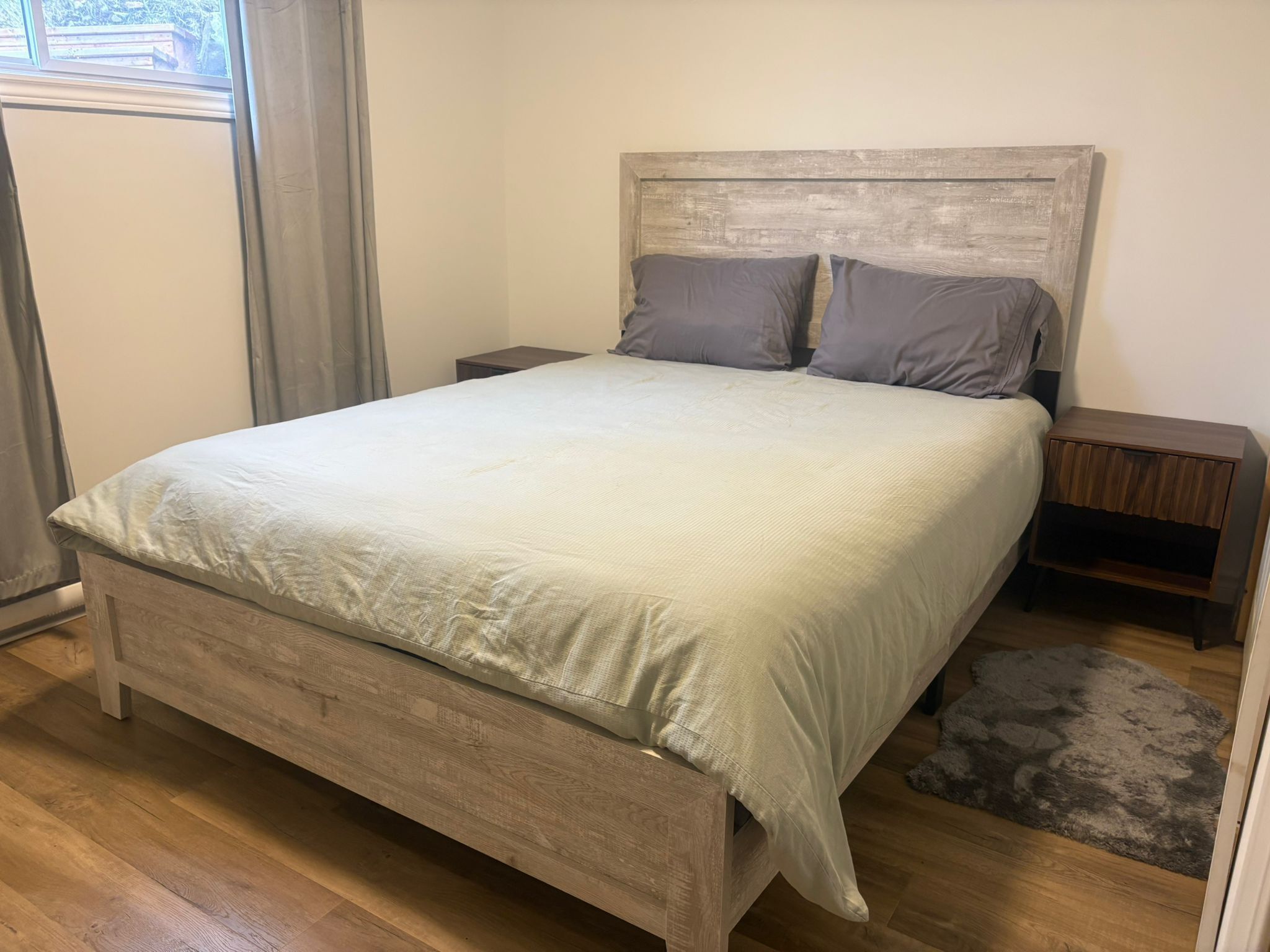 A bedroom with a wooden bed, two nightstands, a grey rug, and a window with curtains.