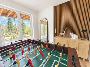 A room with a foosball table, a cabinet with decorative items, and a view of trees through large windows. - Modern Spa Chalet near Lac des Îles · Ski Montcalm 20 min · Pets OK (Entrelacs)