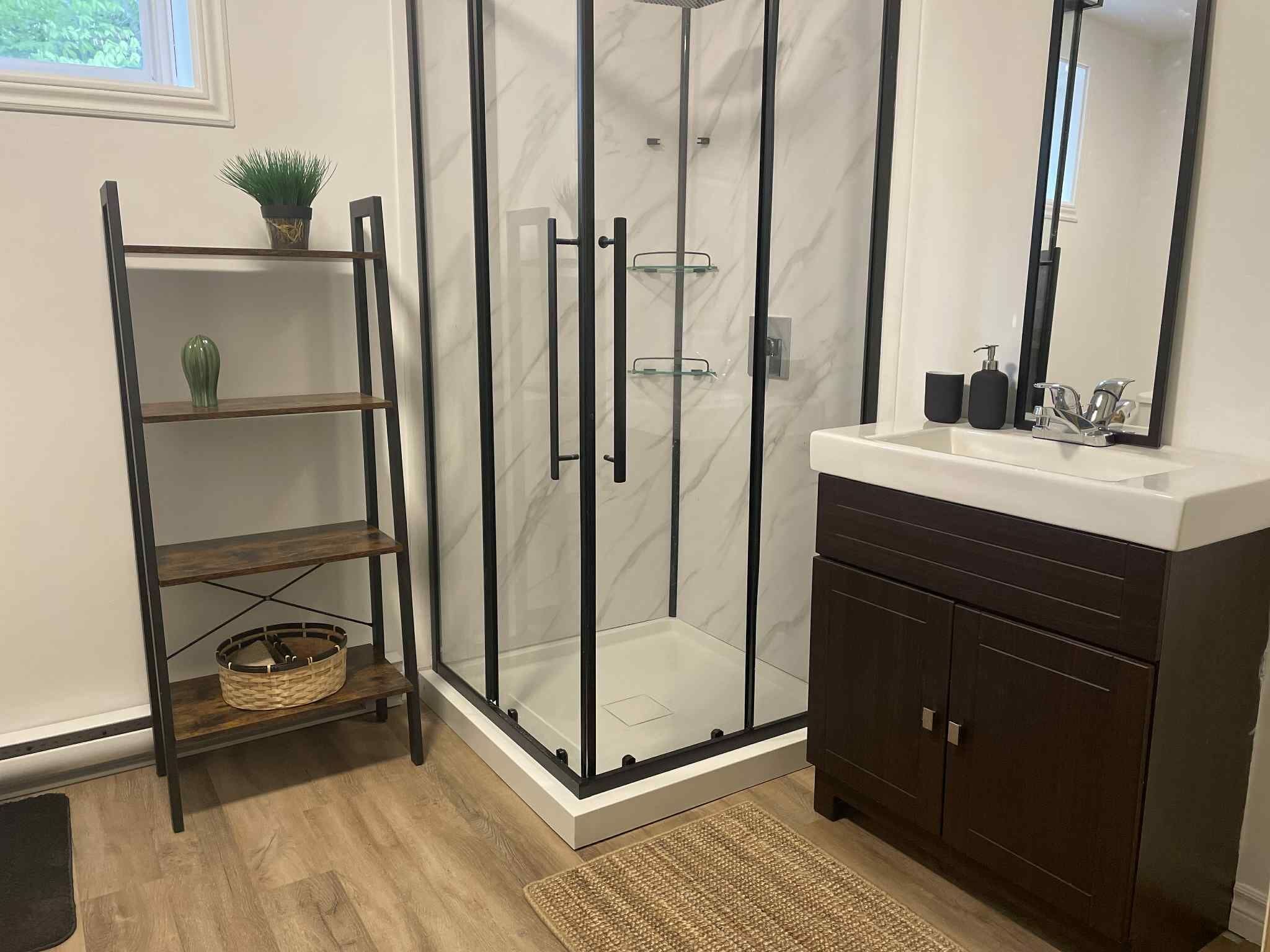 A modern bathroom with a glass-enclosed shower, a wooden shelf with a plant, a dark wooden vanity, and a large mirror.