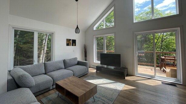 A spacious living room with a grey sectional sofa, a wooden coffee table, and a flat-screen TV. Large windows provide ample natural light and views of the outdoors. - Entire chalet with Spa near Lake & Beach (Entrelacs)