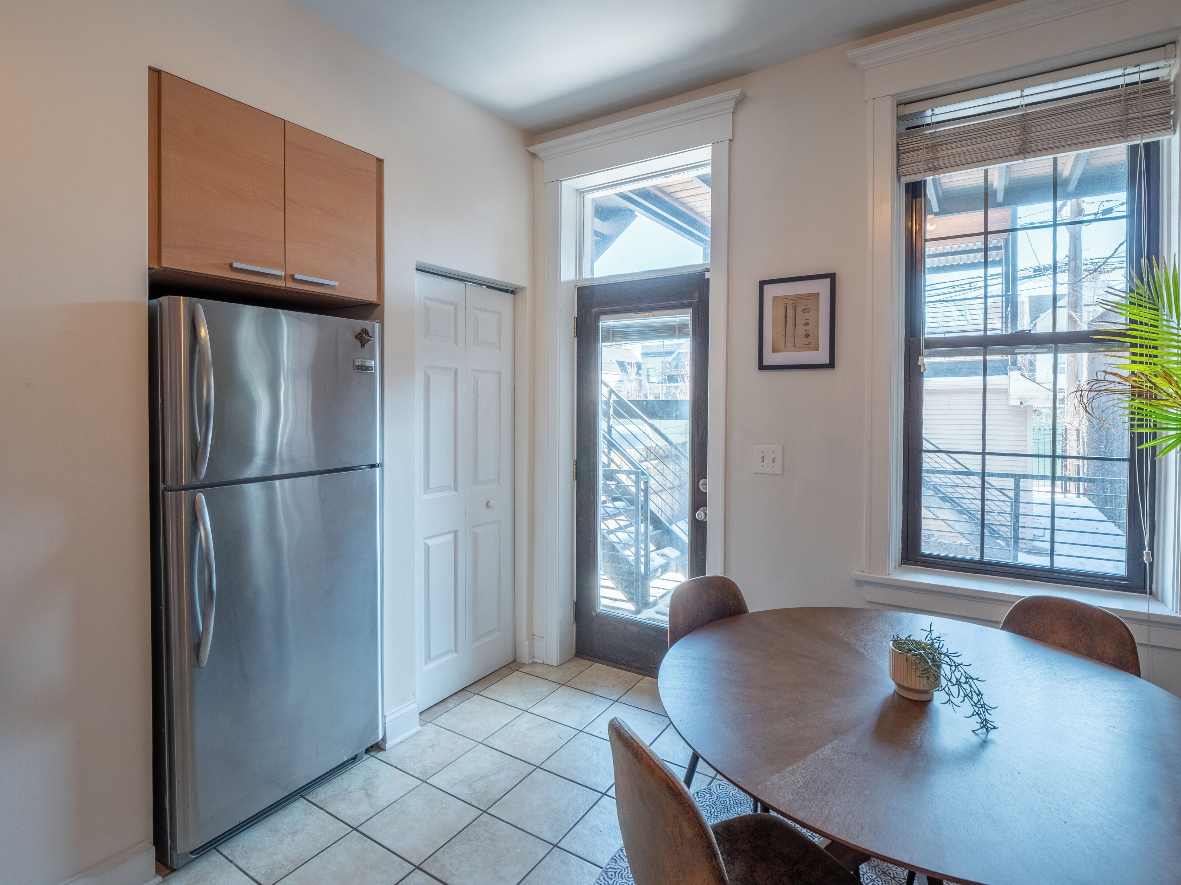 A kitchen with a stainless steel refrigerator, a wooden cabinet, a dining table with chairs, and a window with blinds.