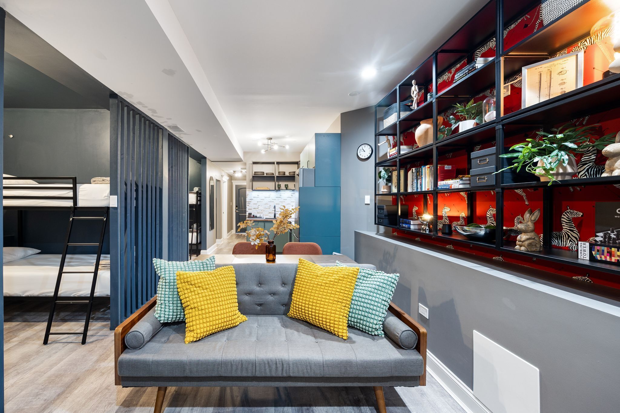 Uma sala de estar moderna com um sofá cinza, almofadas estampadas em amarelo e verde, uma estante com objetos decorativos e uma área de jantar ao fundo.