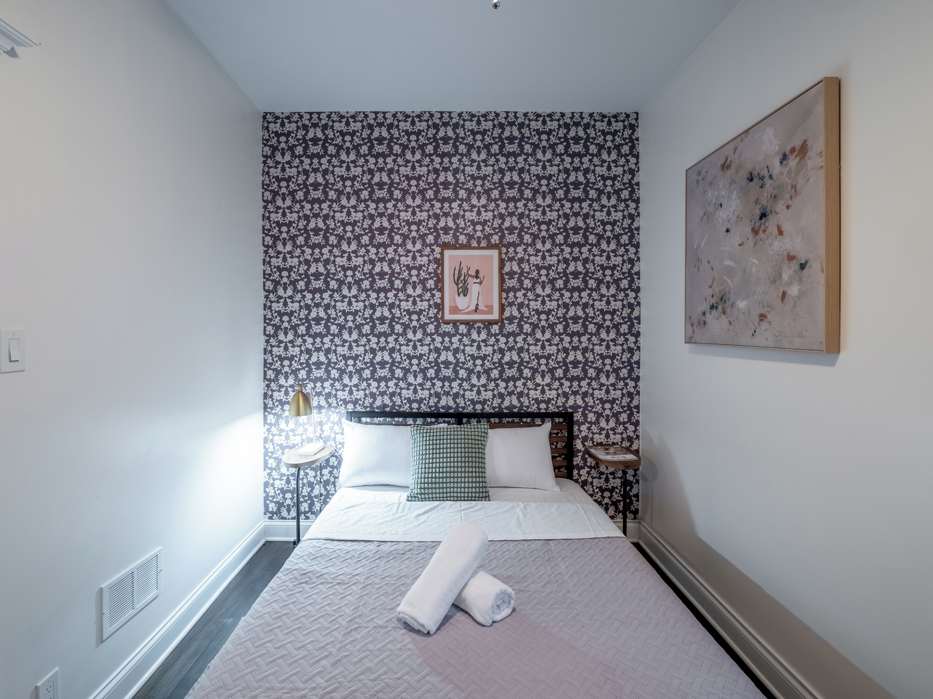 A neatly arranged bedroom with a bed, a bedside table, a wall-mounted artwork, and a patterned wallpaper.