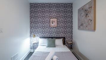 A neatly arranged bedroom with a bed, a bedside table, a wall-mounted artwork, and a patterned wallpaper.