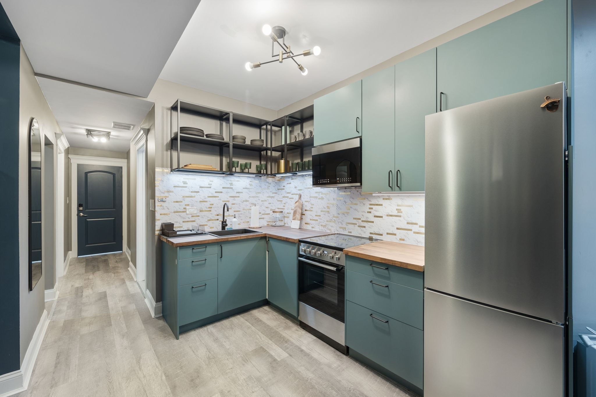 A modern kitchen with teal cabinets, stainless steel appliances, and a herringbone-patterned backsplash.